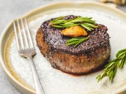 A mouthwatering steak, cooked to medium-rare, with a crispy crust and a juicy center. The steak is garnished with a sprig of rosemary and a clove of roasted garlic.