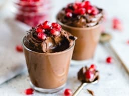A luscious, velvety chocolate mousse, perfectly smooth and creamy, fills two clear glasses. Each glass is adorned with a scattering of vibrant pomegranate seeds.