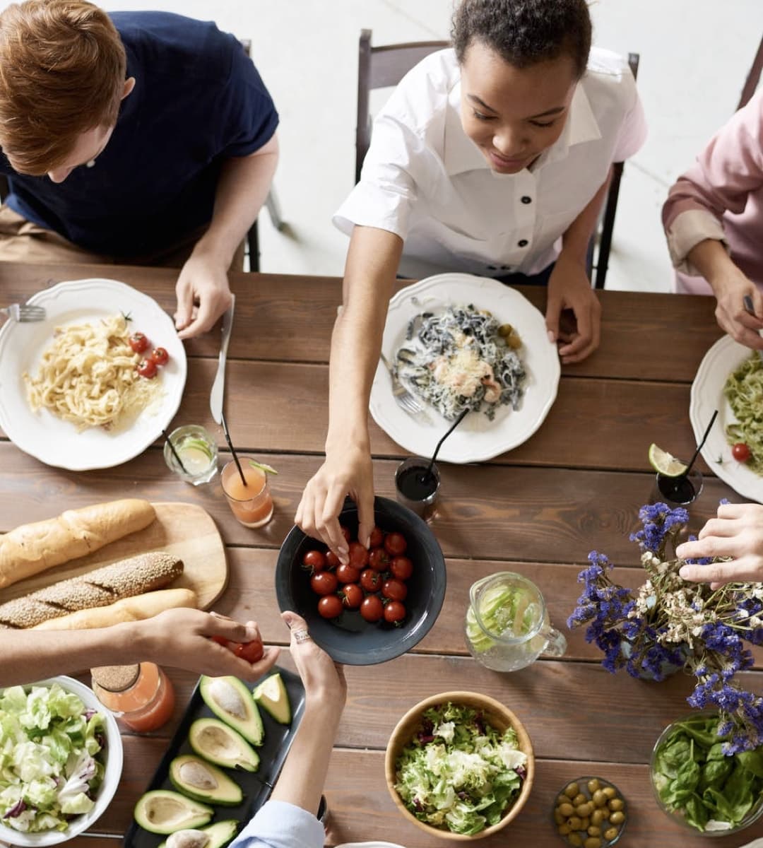 A family seated around a wooden dining table, sharing a meal together. Various dishes are spread across the table, including plates of pasta, a salad bowl, avocados, bread, and a bowl of cherry tomatoes being passed around. The atmosphere is casual and friendly.