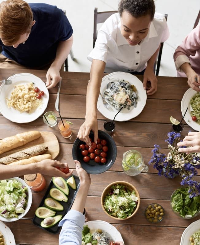 A family seated around a wooden dining table, sharing a meal together. Various dishes are spread across the table, including plates of pasta, a salad bowl, avocados, bread, and a bowl of cherry tomatoes being passed around. The atmosphere is casual and friendly.