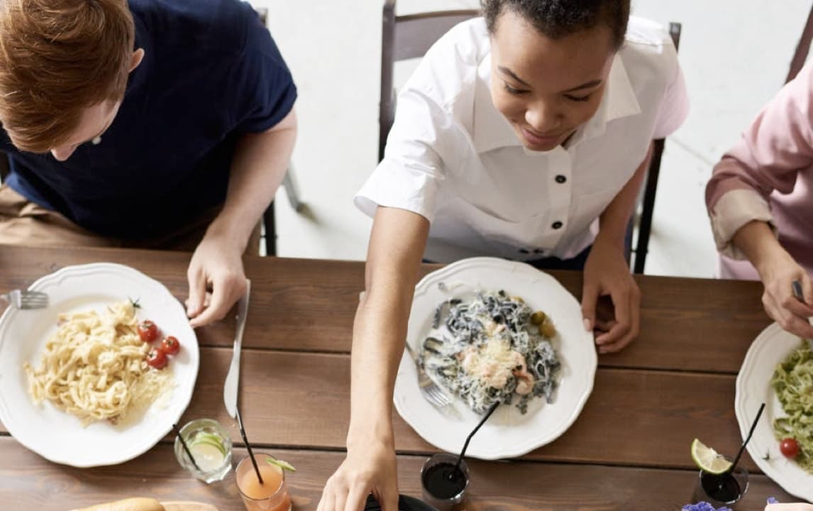 A family seated around a wooden dining table, sharing a meal together. Various dishes are spread across the table, including plates of pasta, a salad bowl, avocados, bread, and a bowl of cherry tomatoes being passed around. The atmosphere is casual and friendly.