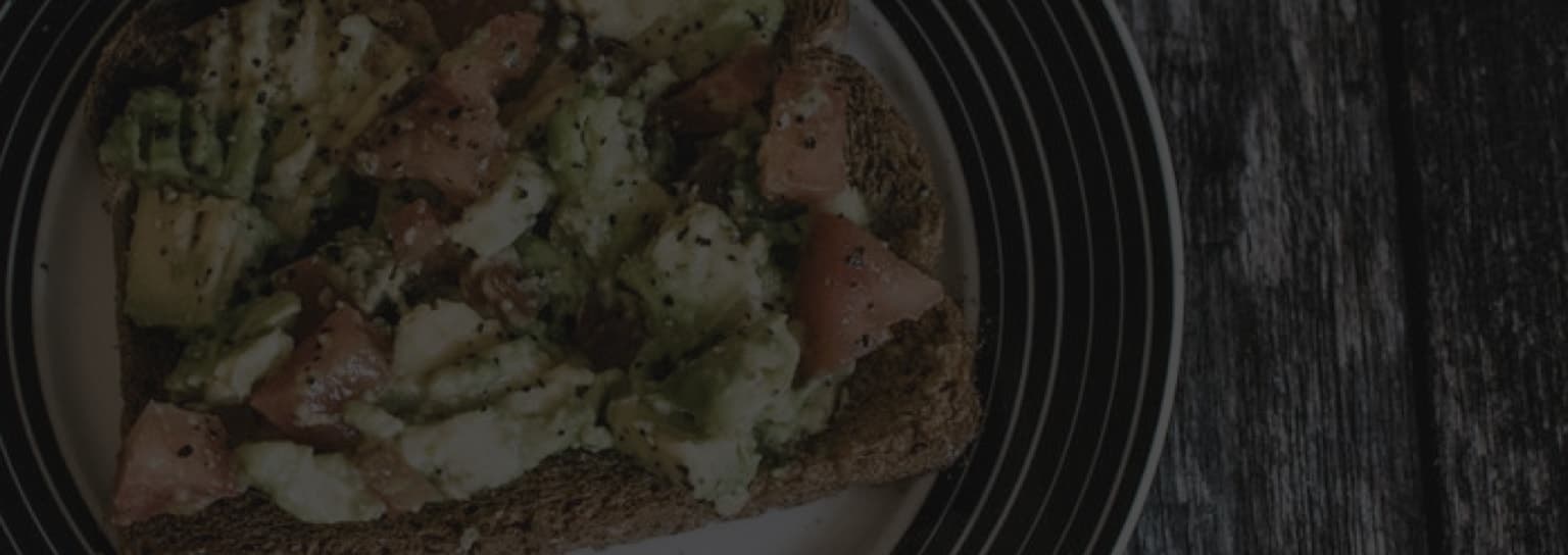 Plate with avocado and tomato on toast, sprinkled with black pepper, on a wooden table background.