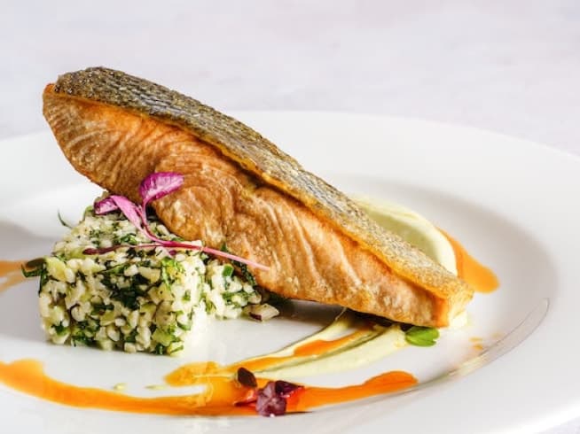 A golden-brown salmon fillet rests atop a mound of fragrant rice, garnished with a sprig of fresh herbs and a drizzle of vibrant orange sauce.