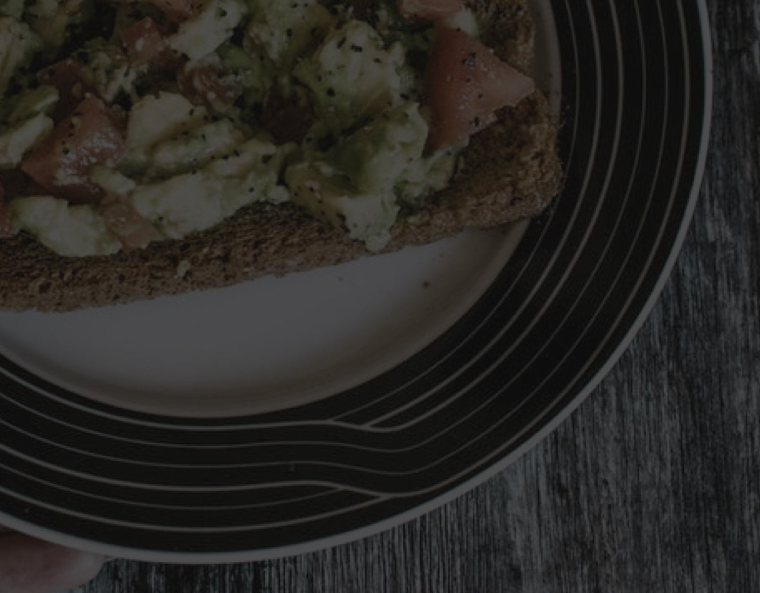 Plate with avocado and tomato on toast, sprinkled with black pepper, on a wooden table background.
